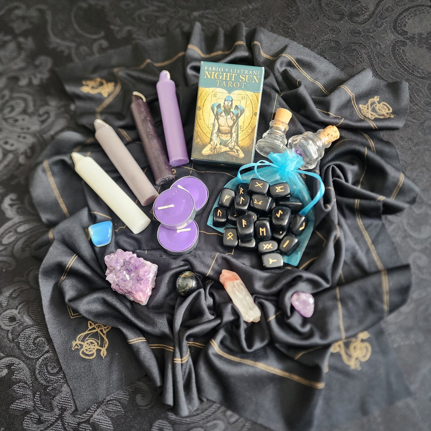 Witchcraft supplies including candles, runes, and crystals on a black cloth with gold patterns.
